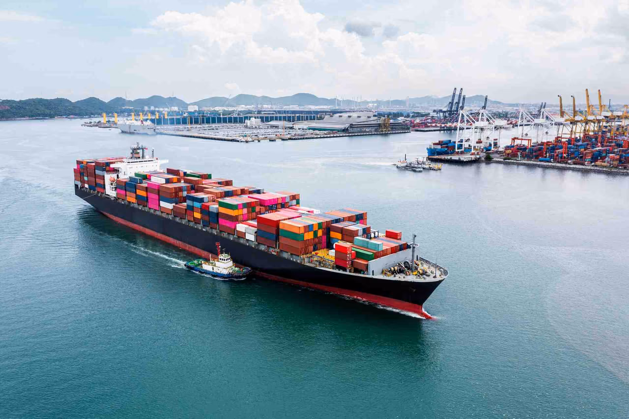 Fully loaded container ship with a tugboat alongside, passing a container port with gantry cranes