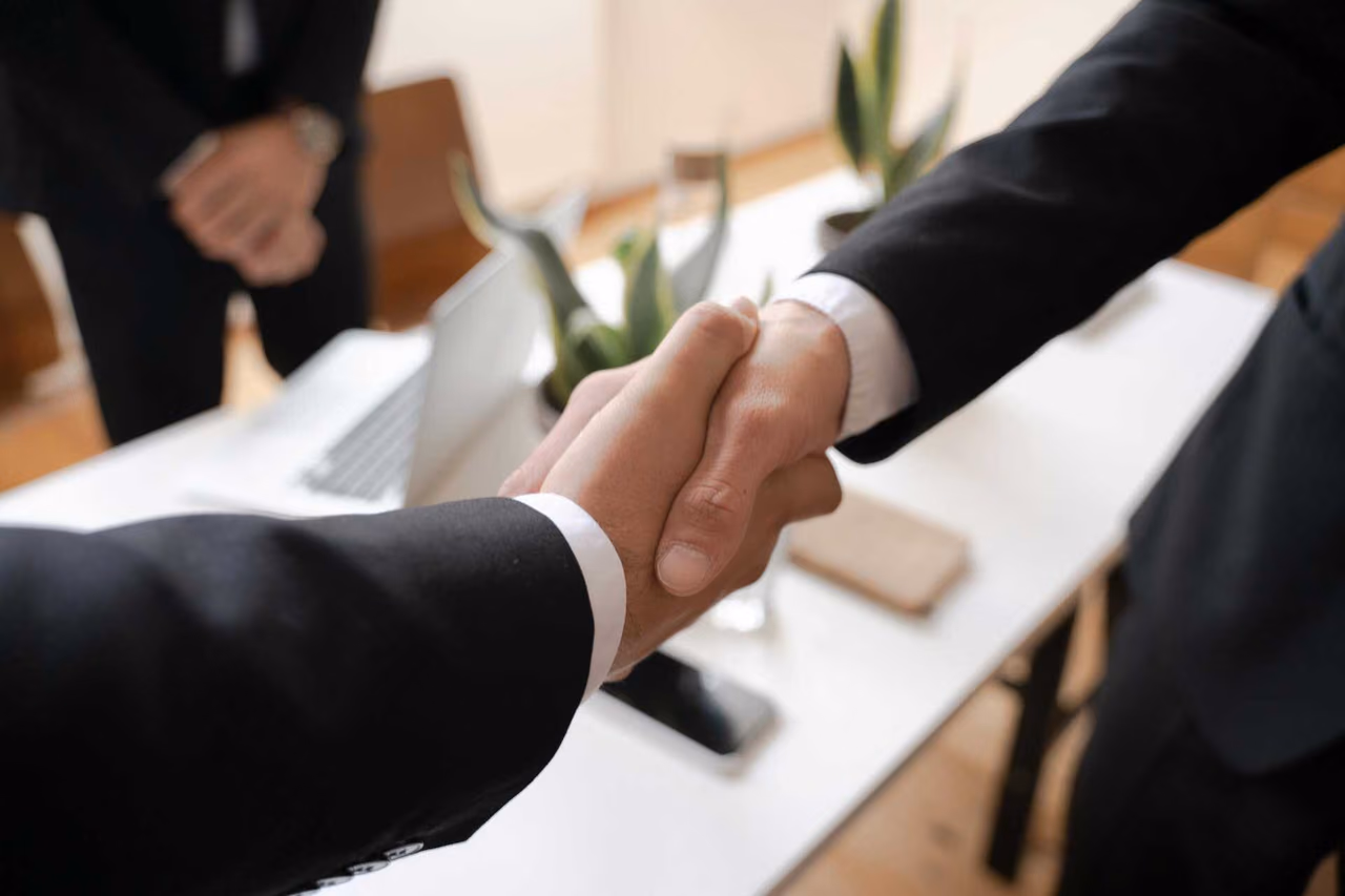 Two people in dark suits shaking hands over a white desk with a laptop and a plant, a third person standing in the background