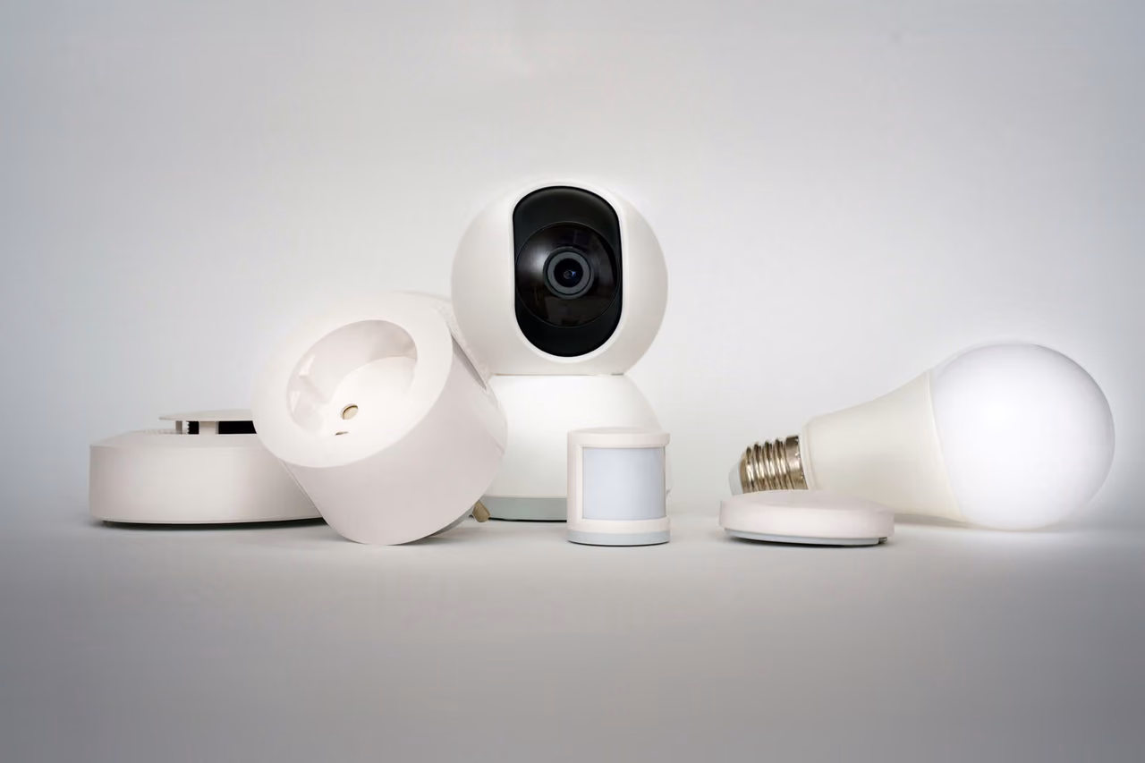 White smart home devices, including a camera, a smart plug, a sensor and a light bulb, on a light grey surface